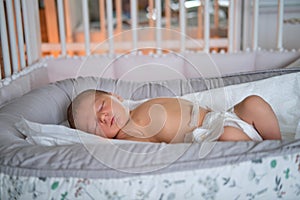 A baby boy sleeps in a cocoon on a cot. A newborn baby in a diaper is lying naked on the bed