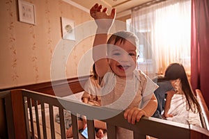 Baby boy smiles at the camera, holding on to his crib. Blurred happy family from behind