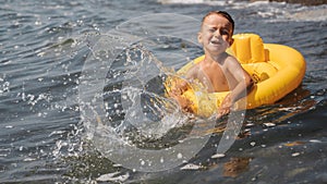 Baby boy splashes water in the sea, floating in a yellow inflatable circle. The kid is having fun and fooling around