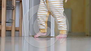 Baby boy taking first steps . Unrecognizable Little child girl walking on white floor at home. Barefoot legs on kitchen.