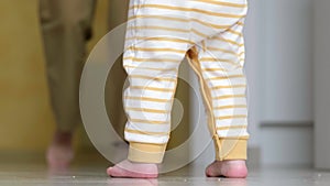 Baby boy taking first steps . Unrecognizable Little child girl walking on white floor at home. Barefoot legs mother and