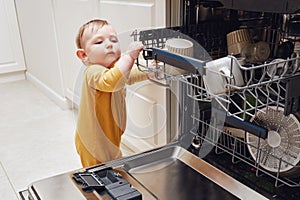 Baby boy toddler is standing by an open dishwasher with dirty dishes i