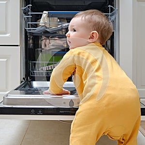 Baby boy toddler is standing by an open dishwasher with dirty dishes i