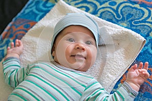 The baby with a cap is lying on the bed and laughing