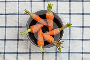 Baby carrots arranged in a circle in black bowl over white table cloth with grid pattern.