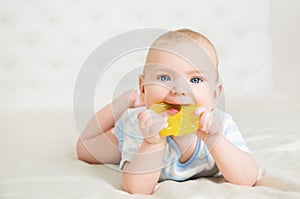 Baby chewing Teether Toy lying down on Stomach in White Bedroom. Child sucking Nibbler and massaging Gums. Babies teething. Cute photo