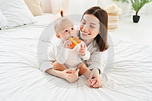 Baby chews on a teething toy while sitting on the bed with mom