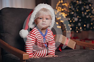 Baby in Christmas pajamas and Santa hat with gift box sitting in armchair at home