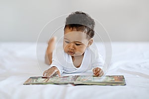 Baby Development Concept. Cute Black Infant Child Lying On Bed With Book