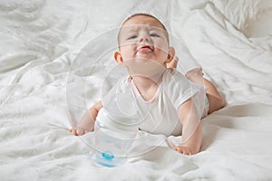 The baby is drooling and teething. He lies on a white bed and pulls his hands to a bottle of water. Cheerless and naughty child