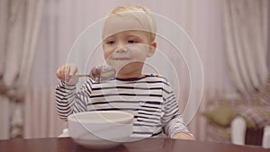 Baby eating. Cheerful baby child eats food itself with spoon. Portrait of beautiful child having breakfast at home. Cute