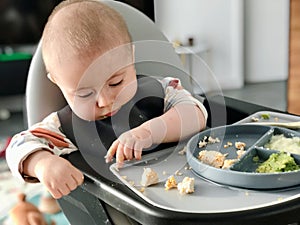 The baby eats independently with his hands while sitting at the feeding table.