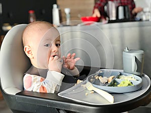 The baby eats independently with his hands while sitting at the feeding table.