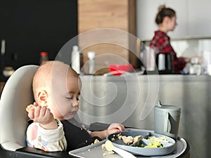 The baby eats independently with his hands while sitting at the feeding table.