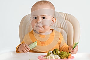 A baby on a feeding chair is eating a cucumber. The concept of proper baby food