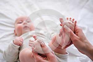 Baby feet in mother hands. Young caucasian woman makes massage for happy infant baby on white bed at home. Babycare photo