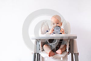 Baby food. little caucasian child drinks water from green plastic cup on highchair on white background. Newborn infant