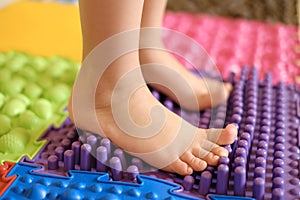 Baby foot standing on an orthopedic massage mat made of puzzles with surfaces of various shapes and tactility.