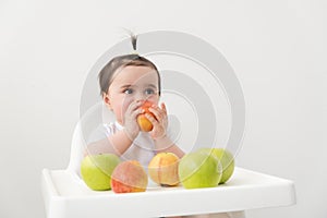 Baby girl in baby chair eating apples and smiling on white background. Baby first solid food