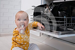 Baby girl biting spoon sits near dishwasher in cozy kitchen