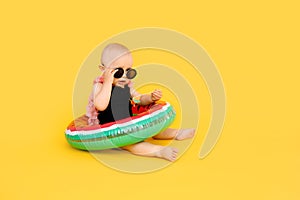 Baby girl in black and pink swimsuit holding watermellon inflatable pool float on yellow background with copy space