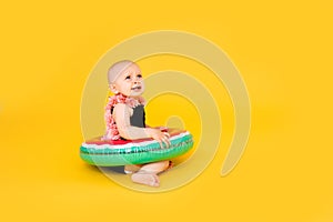 Baby girl in black and pink swimsuit holding watermellon inflatable pool float on yellow background with copy space