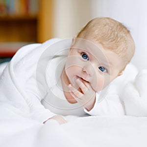 Baby girl with blue eyes wearing white towel or blanket in white sunny bedroom. Newborn child relaxing in bed. Nursery