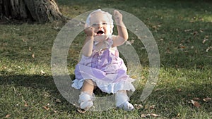 A baby girl in a bonnet sitting on the grass and clapping her hands