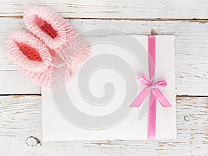 Baby girl booties with a blank card on a white wooden background. Top view. Flat lay.