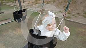 Baby girl with bow on her head is swinging on a swing in the park, little child on the playground