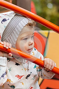 Baby girl boy in a hoodie and hat looks sideways through a metal fence. Caucasian child on a walk holds on to the railing