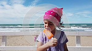 Baby girl child biting popsicle ice cream at beach bar windy sea pink bandana