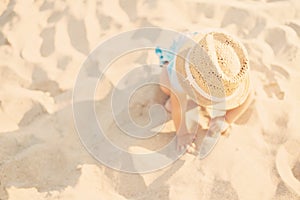 Baby girl child with straw hat and blue dress playing with sand at the beach in summer. Little girl sitting on the shore of the se