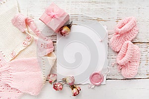 Baby girl clothes, blank card with wax seal and small present box on white wooden background. Flat lay. Owerhad view