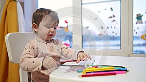 baby girl draws while sitting at a table by the window at home. happy family kid concept. baby daughter learns to draw