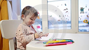 Baby girl draws while sitting at a table by the window at home. Happy family kid concept. Baby daughter learns to draw