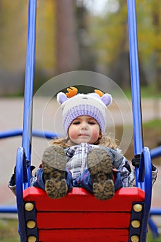 Baby girl in a funny hat swinging on the winter playground, perspective point of view.