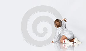 Baby girl in gray bodysuit and socks with red bows. She is creeping on the floor, touching wall, isolated on white. Back view