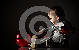 Baby girl in gray boots and sparkling suit. She posing with three red balls, sitting on floor. Twilight black background. Close up