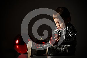 Baby girl in gray boots and sparkling suit. She posing with three red balls, sitting on floor. Twilight black background. Close up