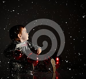 Baby girl in gray boots and sparkling suit. She posing with three red balls, sitting on floor. Twilight black background. Close up