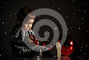 Baby girl in gray boots and sparkling suit. She posing with three red balls, sitting on floor. Twilight black background. Close up