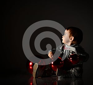 Baby girl in gray boots and sparkling suit. She posing with three red balls, sitting on floor. Twilight black background. Close up