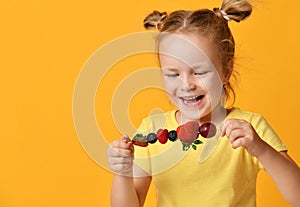 Baby girl kid in blank t-shirt holding smelling biting eating fresh berries dessert with cherry strawberry raspberry on skewer