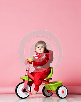 Baby girl kid riding her first bicycle tricycle in red overalls looking up on light pink