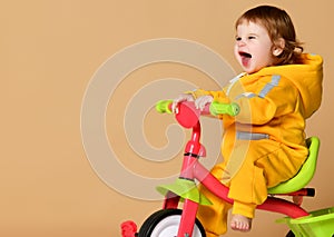 Baby girl kid riding her first bicycle tricycle in warm yellow overalls looking up on light grey