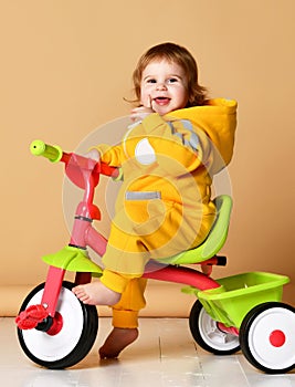 Baby girl kid riding her first bicycle tricycle in warm yellow overalls looking up on light grey