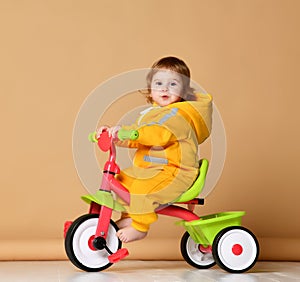 Baby girl kid riding her first bicycle tricycle in warm yellow overalls looking up on light grey