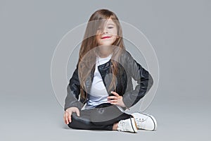 Baby girl with long hair in black leather jacket and leggings is sitting in the studio on grey background