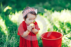 Baby girl in red dress eating pear in summer garden near red bucket